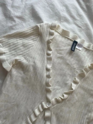 Vit ribbad kofta med volang från H&M - Supersöt vit ribbad kofta från H&M Divided med volangdetaljer längs v-ringningen och en knapp framtill. Kortärmad och i mjukt, stretchigt material som känns skönt mot huden. Perfekt till jeans eller kjol för en fräsch look. Ganska kort också 