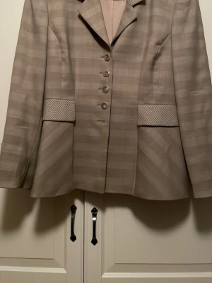 Beige rutig kavaj med knappar - Snygg beige kavaj med rutigt mönster och markerade fickdetaljer. Kavajen har klassisk krage, långa ärmar och stängs med fem knappar framtill. Storlek 42, men upplevs större, snarare 44-46.