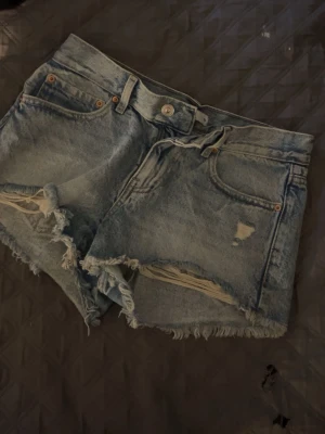 Perfekta jeansshorts med slitningar - Säljer ett par ljusblå denimshorts från Perfect Jeans i storlek 32. De är högmidjade med råa, fransiga kanter och slitna detaljer framtill för en riktigt chill vibe. Klassisk femficksmodell och bälteshällor. Perfekta till sommaren!