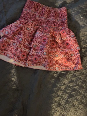 Rosa mönstrad kortkjol med smock - Supersöt kortkjol i rosa med färgglatt mönster och bred smock i midjan. Kjolen har en volangig form och är perfekt för dig som gillar färg och mönster. Lätt och luftig känsla, passar perfekt till varmare dagar.