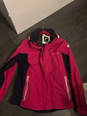 Rosa vindjacka från Five Seasons - Säljer en snygg rosa och marinblå vindjacka från Five Seasons i storlek EU 44. Jackan har dragkedja framtill, två fickor med vita dragkedjor och hög krage. Perfekt för blåsiga dagar och outdoor-äventyr. Materialet är slitstark polyester.