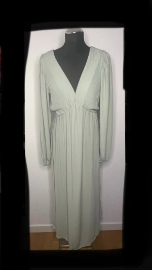 Ljusgrön långklänning med v-ringning - Säljer en ljusgrön långklänning i storlek XS med djup v-ringning och långa ärmar. Klänningen har ett mjukt, flowy tyg och resår i midjan för en snygg siluett. Perfekt för dig som gillar stilrena och enkla plagg med lite extra edge. Aldrig blivit använd