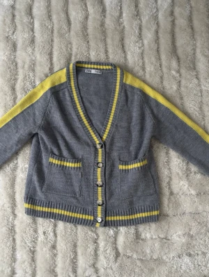 Grå kofta med gula detaljer från ZARA - Grå kofta från ZARA med gula ränder längs ärmar, kanter och fickor. V-ringad med knappar framtill och två fickor. Ribbstickade muddar och mjukt material som passar perfekt till lager på lager under höst och vår.