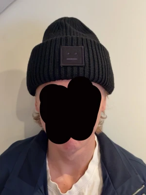 Svart mössa från Acne Studios - Säljer en svart ribbstickad mössa från Acne Studios med deras ikoniska ansiktslogga framtill. Mössan är i ett tjockt, stickat material som håller dig varm och har en bred uppvikt kant. Perfekt accessoar för kyliga dagar.