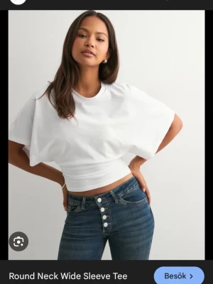 Vit croppad t-shirt med vida ärmar - Snygg vit t-shirt med rund hals och extra vida korta ärmar. Modellen är croppad och sitter tajt i midjan vilket ger en modern och trendig look. Perfekt att matcha med höga jeans för en cool stil.