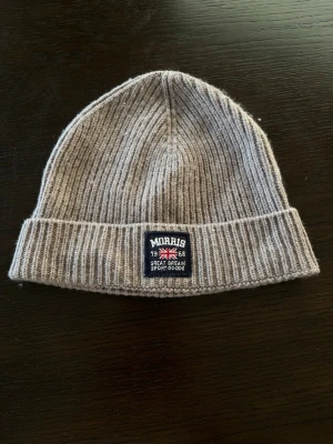 Beige ribbstickad mössa från Morris - Säljer en beige ribbstickad mössa från Morris med patch framtill och snygg uppvikt kant. Mössan är tillverkad i en lyxig mix av 90% merinoull och 10% kashmir, vilket gör den både mjuk och varm. Perfekt accessoar för kyliga dagar.