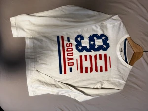 Vit långärmad t-shirt med tryck från Max - Vit långärmad t-shirt från Max med stort tryck på framsidan. Trycket har blå och röda detaljer, texten 'SQUAD' och siffror med stjärnor och ränder. Materialet är mjuk bomull och passformen är normal. Perfekt för dig som gillar sportig och casual stil.