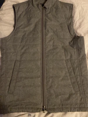 Grå quiltad väst med dragkedja - Säljer en stilren grå quiltad väst med hel dragkedja framtill och två snedställda fickor. Västen har en enkel, rak passform och är fodrad på insidan för extra komfort. Perfekt att slänga över en hoodie eller tröja när det är kyligt ute.