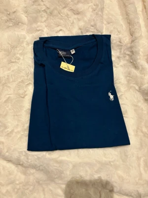 Blå t-shirt från Polo Ralph Lauren - Snygg blå t-shirt från Polo Ralph Lauren med klassisk rund hals och ikoniska vita broderade loggan på bröstet. Tillverkad i mjuk bomull som känns skön mot huden. Perfekt till jeans eller shorts för en clean look.