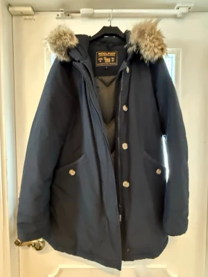 Woolrich vinterjacka L – äkta & varm - Säljer en klassisk och varm Woolrich parkas i mörkblått. En tidlös modell som håller värmen riktigt bra under kalla vinterdagar.  ✨ Detaljer: 	•	Storlek: L 	•	Färg: Mörkblå 	•	Varm fodrad vinterjacka 	•	Huva med pälsdetalj 	•	Stängning med dragkedja + knappar 	•	Två stora fickor framtill 	•	Original Woolrich-detaljer och märken 	•	Material: Ramar Cloth (60% bomull / 40% nylon)