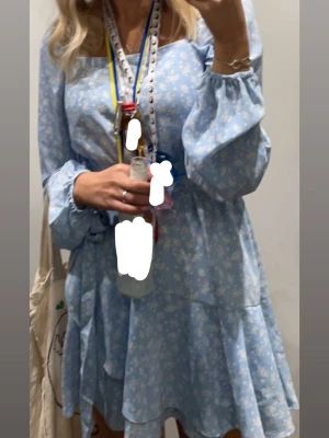 Blå blommig kortklänning med volang - Supersöt ljusblå kortklänning med vita små blommor, långa ärmar och volangdetalj nertill. Klänningen har fyrkantig ringning och smockad rygg för extra bekväm passform. 