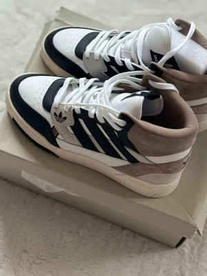 Adidas sneakers i vitt, svart och beige - Säljer ett par Adidas sneakers med högt skaft i vitt, svart och beige. Skorna har klassiska tre ränder på sidan, snörning och detaljer i mocka och skinn. Perfekta för dig som vill ha en clean och sportig look med retrovibbar.