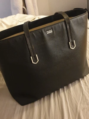 Svart handväska från Hugo Boss - Snygg svart handväska från Hugo Boss med stilren design och silverfärgade detaljer. Väskan har två långa handtag, strukturerad yta och rymligt beige innerfoder med flera fack och dragkedja. Perfekt för dig som vill ha en klassisk och praktisk väska.