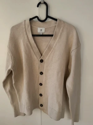 Beige kofta från H&M - Säljer en beige kofta från H&M i relaxed fit. Koftan är stickad i 100% bomull, har långa ärmar och knäpps med fem mörka knappar framtill. Perfekt för lager på lager och enkel att styla till olika outfits. Storlek S passar Xs