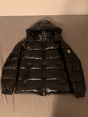 Svart Moncler Maya pufferjacka - Säljer en svart, glansig pufferjacka från Moncler med huva och dragkedja framtill. Jackan har två snedställda fickor med dragkedja, en ficka med Moncler-logga på ärmen och quiltad design. Insidan har ett coolt Moncler-serietryck. Perfekt för kalla dagar.