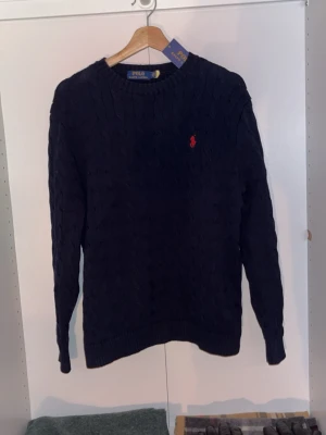 Mörkblå stickad tröja Polo Ralph Lauren - Säljer en mörkblå kabelstickad tröja från Polo Ralph Lauren i storlek M. Tröjan har rund hals, lång ärm och klassisk röd broderad logga på bröstet. Materialet är mjuk bomull och passformen är normal. Perfekt för dig som gillar stilrena och tidlösa plagg.