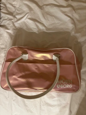 Rosa axelväska från Björn Borg - Säljer en retroinspirerad rosa axelväska från Björn Borg med vita detaljer och handtag. Väskan har ett stilrent tryck med loggan på framsidan och dragkedja upptill. Materialet är glansigt syntet som ger en cool look.