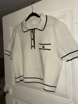 Vit topp med krage från H&M - Säljer en vit topp med krage från H&M med svarta kontrastkanter och guldknappar framtill. Toppen har korta ärmar, krage och en dekorativ ficka på bröstet. Perfekt för en preppy look och enkel att styla till jeans eller kjol.