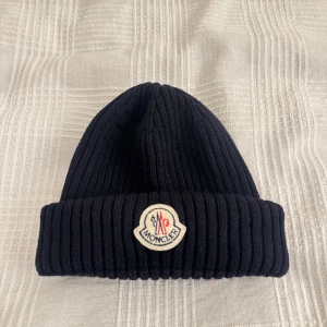 Marinblå ribbad Moncler mössa - Säljer en äkta marinblå ribbstickad mössa från Moncler med klassisk logga framtill. Mössan är i tjockt, mjukt material och har vikt kant för extra stilpoäng. Perfekt accessoar för kalla dagar.