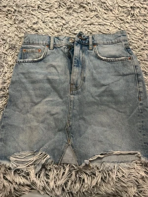 Ljusblå jeanskjol med fransig kant - Säljer en ljusblå jeanskjol från Premium Denim med rå, fransig kant och slitningar längst ner. Kjolen har klassisk femficksdesign, bälteshällor och knappgylf. Perfekt för dig som gillar en avslappnad och trendig stil.