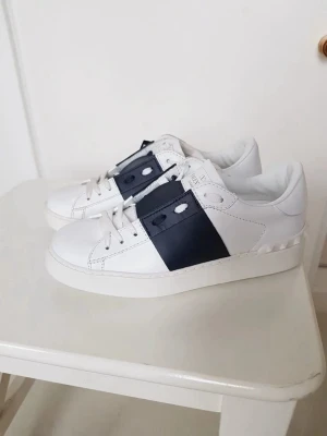 Valentino Garavani Snörade sneakers vit/navy  - Säljer ett par Valentino Garavani laced sneakers i vitt skinn med bred navy blue läderdetalj över mitten. Klassisk rund tå, vita snören och ikoniska nitar på hälen. Kommer med originalkartong och dustbag. Perfekta för dig som vill ha en clean men lyxig look.
