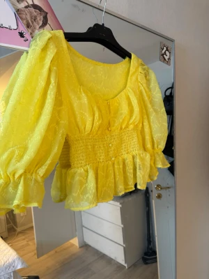 Gul blus med volanger och puffärm - Säljer en superfin gul blus med transparent tyg, puffiga ärmar och bred smock i midjan. Blusen har volangkant nedtill och vid ärmslut, samt små dekorativa knappar framtill. Perfekt för dig som vill ha något färgglatt och trendigt till sommaren!