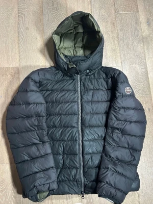 Svart pufferjacka från Colmar - Säljer en svart pufferjacka från Colmar med huva och grönt innerfoder. Jackan har quiltad design, dragkedja framtill och långärmade ärmar. Perfekt för kalla dagar och riktigt skön att bära.