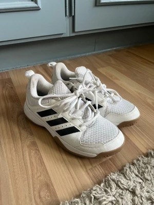 Vita Adidas sneakers - Säljer ett par vita Adidas sneakers med klassiska svarta ränder på sidorna. Skorna har snörning, meshdetaljer och en gummisula i beige. Perfekta för dig som gillar sportig stil och vill ha bekväma skor till vardagen. 