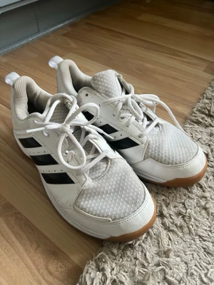 Vita adidas sneakers - Säljer ett par vita adidas sneakers med klassiska svarta ränder på sidorna. Skorna har snörning, mesh-överdel och gummisula i ljusbrun ton. Perfekt för dig som gillar sportig stil och vill ha bekväma skor till vardagen. Storlek 42 