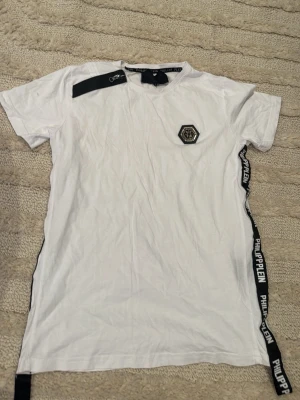 Vit Philipp Plein t-shirt med logoband - Säljer en vit t-shirt från Philipp Plein med svart logoband längs båda sidorna och axeln. T-shirten har ett svart emblem på bröstet och rund hals. Materialet är mjuk bomull och modellen är normal passform. 