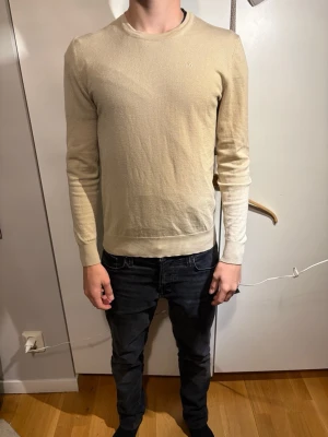 Beige merino ull  tröja från J.Lindeberg - Funkar för xs Snygg beige stickad tröja från J.Lindeberg med diskret logga på bröstet. Tröjan har rund hals, ribbade muddar vid ärmslut och nederkant samt långärmad passform. Perfekt för dig som gillar stilrena och enkla plagg.