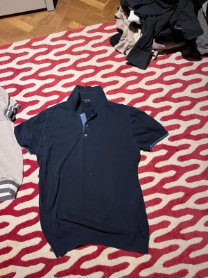 Mörkblå pikétröja med krage - Säljer en mörkblå pikétröja från Massimo Dutti med klassisk krage och knappar framtill. Tröjan har korta ärmar och ribbade muddar vid ärmslut och nederkant. Perfekt till sommaren och enkel att matcha med jeans eller shorts.