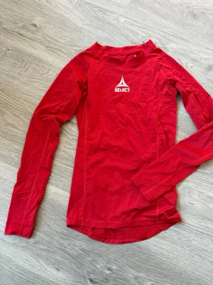 Röd långärmad träningströja Select 14/16 - Säljer en röd långärmad träningströja från Select med vit logga på bröstet. Tröjan är tillverkad i ett lätt och stretchigt syntetmaterial som passar perfekt för sport och aktivitet. Snygg passform och rund hals.