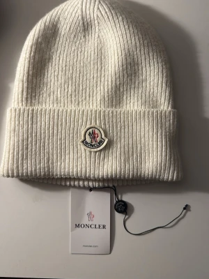 Ingen väst på bilderna - vit stickad mössa från Moncler med uppvikt kant och broderad Moncler-logga framtill. Mössan har en ribbad struktur och medföljande tagg