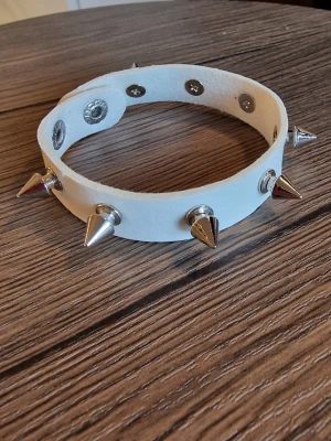 Vit punk choker med silverfärgade nitar - Armbaand punk. Cool vit choker med flera silverfärgade konformade nitar och tryckknappar. 24 cm långt. Bredd 1,5 cm. Armbandet är tillverkat i syntetmaterial Pu läder och har en edgy vibe som passar perfekt till streetwear eller alternativa stilar.