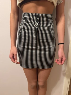 Randig kortkjol från Divided H&M - Säljer en svartgrå randig , högmidjad kortkjol från H&M. Med  tre knappar framtill, fickor både fram och bak samt en snygg detalj med liten v-formad öppning bak i midjan. Aldrig använd i storlek xs/34