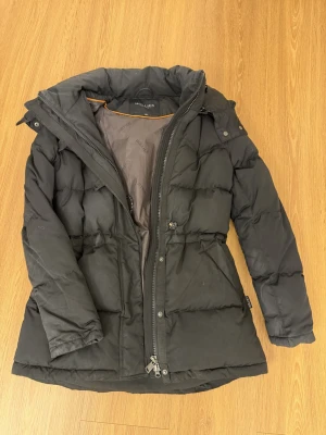 Svart pufferjacka från Hollies, strl 36 - Svart pufferjacka från Hollies med hög krage och dragkedja framtill. Jackan har quiltad design, justerbar midja med knapp och snygga detaljer som logotyp på nederkanten. Perfekt för kalla dagar och enkel att matcha med din stil. Priset kan diskuteras vid snabb affär.