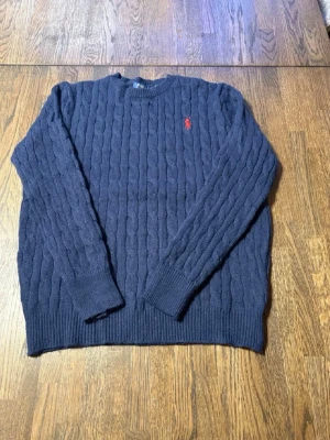 Mörkblå stickad tröja Polo Ralph Lauren - Mörkblå stickad tröja från Polo Ralph Lauren i storlek L. Storlek stämmer inte överens med passformen och skulle anse att den mer är M. Nästan oanvänd och har inga defekter eller hål. Klassisk kabelstickad design med rund hals och rött broderat logomärke på bröstet. Perfekt för dig som gillar stilrena och tidlösa plagg. Fraktar på 1-2 dagar!