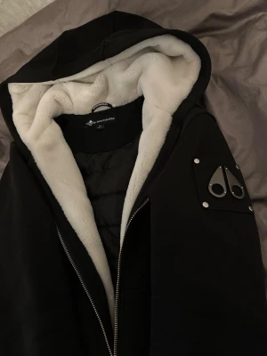 Svart pufferjacka Moose Knuckles - Svart pufferjacka från Moose Knuckles. Jackan har en snygg metallpatch på ärmen, quiltad insida och grov dragkedja framtill. Perfekt för kalla dagar och har en cool streetstil.( Barnstorlek ) L