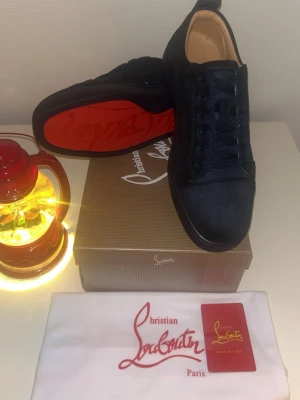 Svarta sneakers från Christian Louboutin - Säljer ett par svarta sneakers från Christian Louboutin med klassisk röd sula och rund tå. Skorna har snörning och är tillverkade i mocka med snygga detaljer. Perfekt för dig som vill ha något lyxigt och stilrent i garderoben.