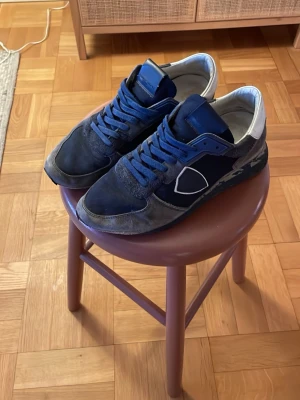 Phillipe Model skor - Snygga blå sneakers från Philippe Model med mörkblå och grå toner, vita detaljer och klassisk logga på sidan. Skorna har snörning, rund tå och platt sula. Materialmix av mocka och syntet ger en cool look. Perfekta för dig som gillar streetstyle och vill sticka ut.