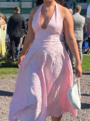 Ljusrosa långklänning med halterneck - Superfin ljusrosa långklänning med halterneck och djup v-ringning. Klänningen har ett blommigt mönster i tyget och är tillverkad i ett glansigt material som ger en lyxig känsla. Kjolen är utsvängd och ger en elegant siluett. Perfekt för speciella tillfällen!