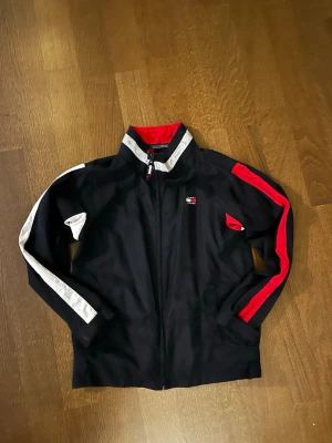  Tommy Hilfiger ungdom XL Vindjacka - Svart vindjacka från Tommy Hilfiger med röda och vita detaljer på ärmarna och insidan. Jackan har dragkedja framtill, ståkrage med vit rand och meshfoder i rött. Klassisk logga på bröstet och snygga kontrastpaneler längs ärmarna.
