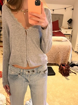 Grå  kofta med spets kant - Säljer en grå  kofta med lång ärm och v-ringad hals. Koftan har spets kant runt halsen och knäppning med små knappar framtill. Mjuk stickad känsla, perfekt för lager på lager och chill stil.