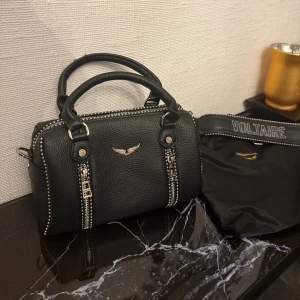 Zadig Voltaire - Cool svart handväska från Zadig & Voltaire i skinn med silverfärgade detaljer och dragkedjor. Väskan har två handtag, avtagbar axelrem med VOLTAIRE-tryck och dekorativa nitar längs kanterna. Perfekt storlek för det viktigaste och snygg design med logga framtill.