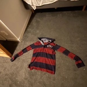 Randig långärmad skjorta från GANT - Säljer en snygg långärmad skjorta från GANT med breda marinblå och röda ränder. Skjortan har klassisk krage, knappar vid halsen och tryckt logga på bröstet. Perfekt för dig som gillar sportig och avslappnad stil.