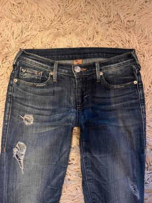 Blå bootcut jeans från True Religion - Snygga blå jeans från True Religion med bootcut passform och låg midja. Jeansen har slitna detaljer och klassiska fickor med den ikoniska hästskosömmen bak. Materialet är robust denim och färgen är mörkblå med tvättade partier. Midjemåttet:37 inerbennslöbgden:84