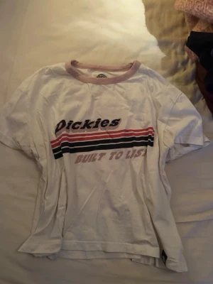 Vit Dickies t-shirt med tryck - Säljer en vit t-shirt från Dickies i storlek small. T-shirten har korta ärmar, rund hals med ljusrosa kant och coolt tryck med texten 'Dickies Built to List' samt ränder i rosa, svart och rött över bröstet. Perfekt för dig som gillar streetwear och enkel stil.