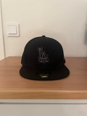 Svart LA 59FIFTY keps från New Era - Säljer en svart LA-keps från New Era, modell 59FIFTY. Kepsen har en platt skärm och broderad LA-logga framtill i mörkgrått. På sidan finns New Era-loggan i vitt och MLB-logga baktill. Insidan har märkning och baseball-detalj. Perfekt för dig som gillar streetwear.