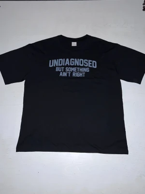 ”Undiagnosed but something ain’t right” t-shirt - En svart t-shirt med ett ironiskt och humoristiskt texttryck. Texten “Undiagnosed but something ain’t right” är tryckt i diskret grå/blå ton som ger ett stilrent men uttrycksfullt intryck. Passar perfekt för dig som gillar självdistans och humor i vardagen. Tröjan är i nytt skick och bekväm att bära.